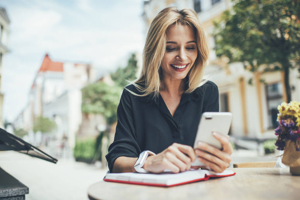 Cheerful female blogger using textbook for planning publication posting sitting in sidewalk and reading notification from social networks, happy hipster girl watching streams video on cellphone - Φωτογραφία, εικόνα