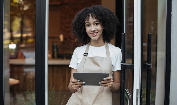 Millennial ιδιοκτήτης του σύγχρονου καφέ αφροαμερικάνικη κυρία με ποδιά κατέχει ψηφιακή ταμπλέτα και στέκεται κοντά στην μπροστινή πόρτα - Φωτογραφία, εικόνα