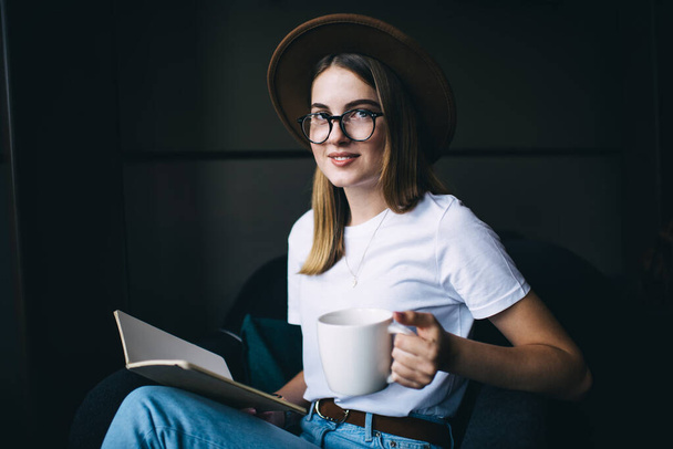 Positive smiling young lady in eyeglasses and trendy hat sitting in comfortable armchair with notebook and mug of hot drink while looking at camera - Photo, Image