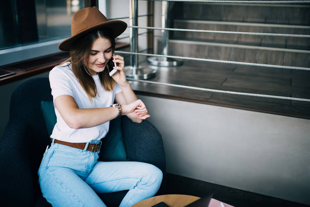High angle of young female freelancer wearing casual clothes and trendy hat making appointment on mobile phone and checking time while smiling and working remotely in cafe - Foto, Imagem