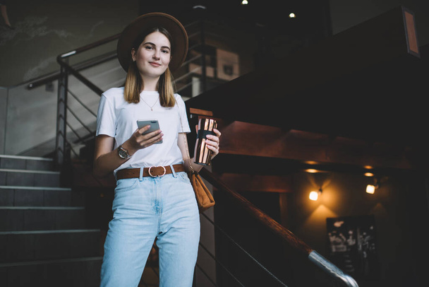 Half length portrait of beautiful trendy dressed female 20s holding thermo cup and smartphone, caucasian attractive hipster girl in stylish apparel looking at camera using mobile phone for blogging - Fotografie, Obrázek