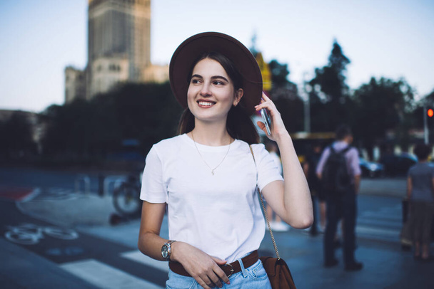 Happy trendy female in stylish hat and handbag with smartphone in hand standing on sidewalk and looking away in city street on summer day - Фото, изображение