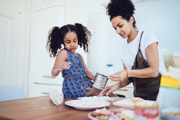 African American mother in white shirt and apron taking flour from daughter in blue dress holding cup in kitchen while cooking together - Foto, afbeelding