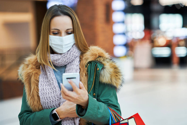 Portrait of adult woman shopping in mall using smartphone wearing a mask - Valokuva, kuva