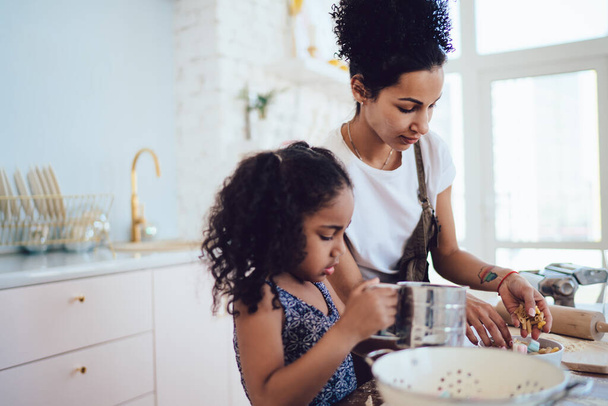 Crop young African American woman and little girl with sifter spending time together and preparing dough standing at table in light kitchen - Foto, afbeelding