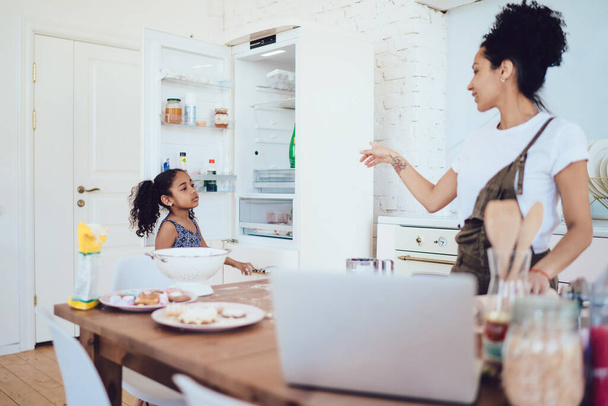 African American mother scolding sad daughter with curly hair for opening fridge while standing in kitchen of modern studio apartment - Foto, imagen