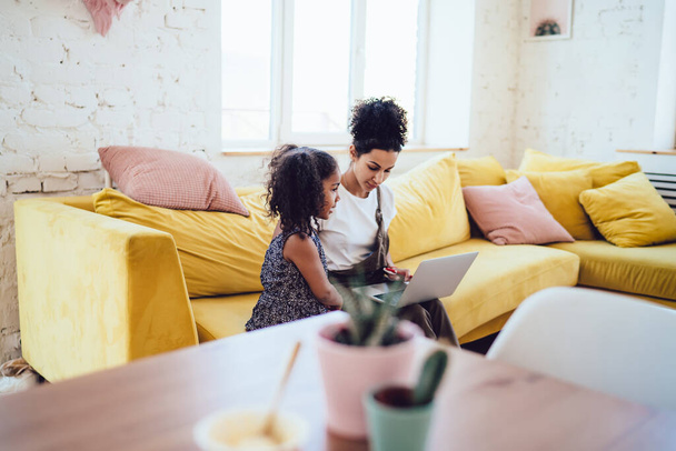 Pretty African American lady with hair bun sitting on soft and comfortable couch near adorable little girl and browsing laptop - Фото, изображение