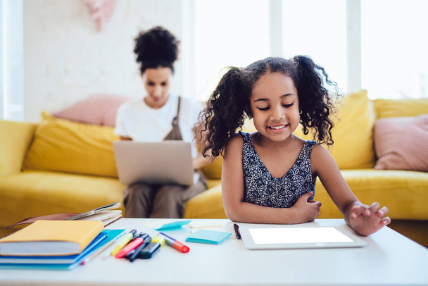 Радісна чорна дитина з посмішкою сидить перед столом з нагадуваннями та маркерами та планшетом для перегляду з порожнім екраном під час роботи з ноутбуком
 - Фото, зображення