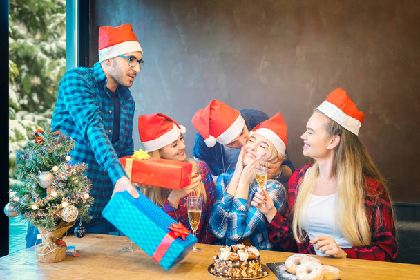 Gruppe von Freunden mit Nikolausmützen feiert Weihnachten, indem sie Geschenke verteilt und zu Hause Champagner trinkt - Foto, Bild