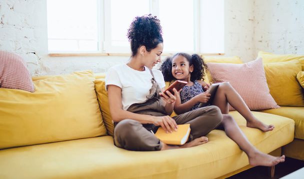 Smiling young black woman holding smartphone and book speaking with happy little daughter with tablet while sitting on yellow sofa in light living room - Φωτογραφία, εικόνα