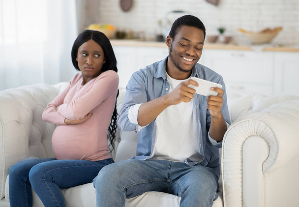 Adicción al juego móvil. Joven hombre negro jugando videojuego en el teléfono inteligente y descuidando a su esposa expectante en casa - Foto, imagen
