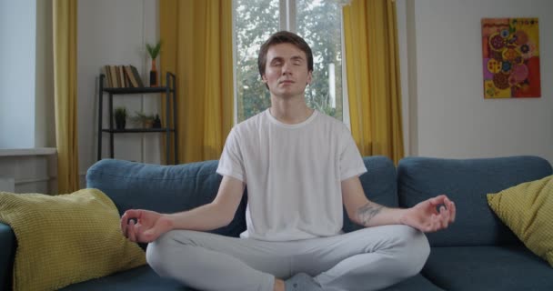Első látásra a fiatalember meditál egyedül otthon természetes fény lassított felvétel. Vonzó személy csukott szemmel ül a kanapén pihentető forgalmas hét szoba háttér. Stresszoldó gyakorlat életmód - Felvétel, videó