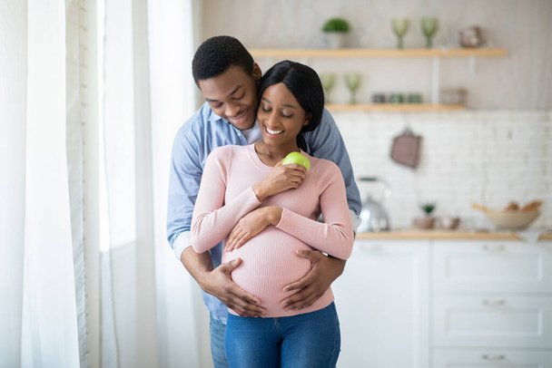Красивая беременная женщина с яблоком и ее парень обнимается в помещении - Фото, изображение