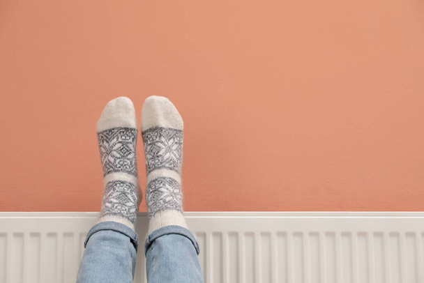 Woman warming legs on heating radiator near color wall, closeup. Space for text - Zdjęcie, obraz
