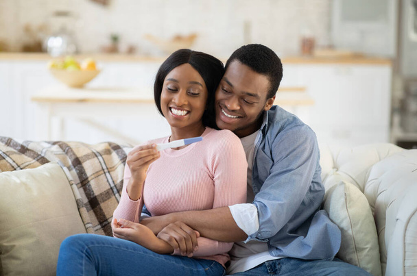 Beautiful black couple holding positive pregnancy test and hugging on sofa at home - Foto, immagini