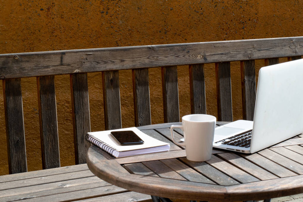 Concepto de trabajar desde casa al aire libre. Portátil, taza de café, portátil y teléfono móvil en la mesa de café de madera frente al banco de madera en el patio. - Foto, imagen