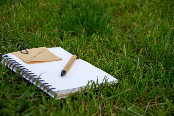 Concept of sustainability with notebook and pen made of recyclable materials and a note with the word Sustainable written on it. Book in the grass outdoors. - Foto, afbeelding