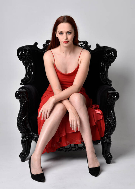 full length portrait of pretty woman wearing red sexy dress. Sitting pose on a black ornate armchair, against a studio background. - Фото, изображение