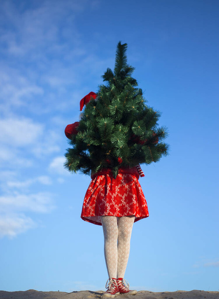 クリスマスのコンセプト。休日の準備。明るい赤いドレスの女の子と青い空に対するサンタの帽子は彼女の人工的なクリスマスツリーの前に保持しています - 写真・画像