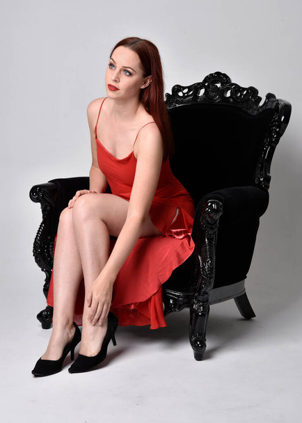 full length portrait of pretty woman wearing red sexy dress. Sitting pose on a black ornate armchair, against a studio background. - Photo, Image