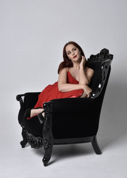full length portrait of pretty woman wearing red sexy dress. Sitting pose on a black ornate armchair, against a studio background. - Photo, Image