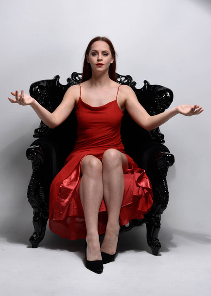 pełnowymiarowy portret pięknej kobiety w czerwonej seksownej sukience. Siedząc na czarnym, ozdobnym fotelu, na tle studia. - Zdjęcie, obraz