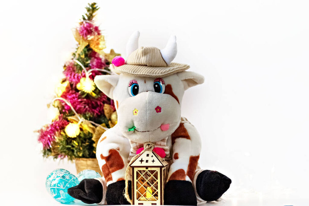 White soft toy bull on a light background with a Christmas tree and garlands, New Year's card. Chinese Year of the Ox, zodiac symbol 2021. - Photo, Image