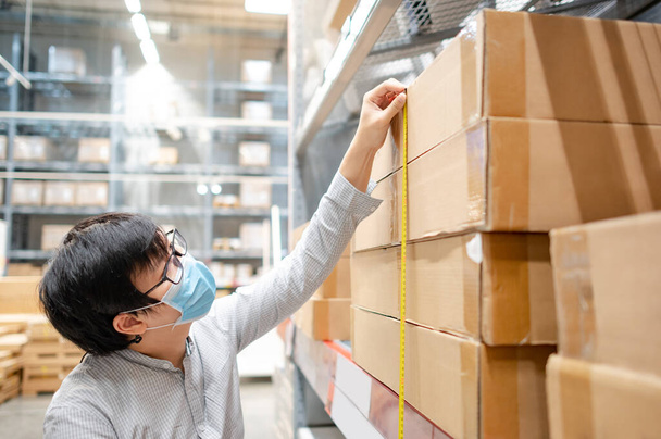 Азиатский работник носит маску с помощью рулетки для измерения размеров продукта в картонной коробке. Контроль качества на складе. - Фото, изображение