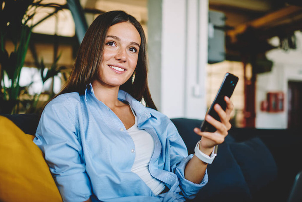 Young cheerful female teenager sitting indoors and holding modern mobile phone, Caucasian smiling woman installing multimedia application on cellular device connecting to 4g internet on leisure - Фото, изображение