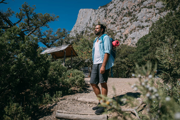 A young man - a tourist with a backpack admires the mountains. Handsome guy with tourist equipment stands on the path among the trees against the background of high rocks and blue sky - Photo, Image