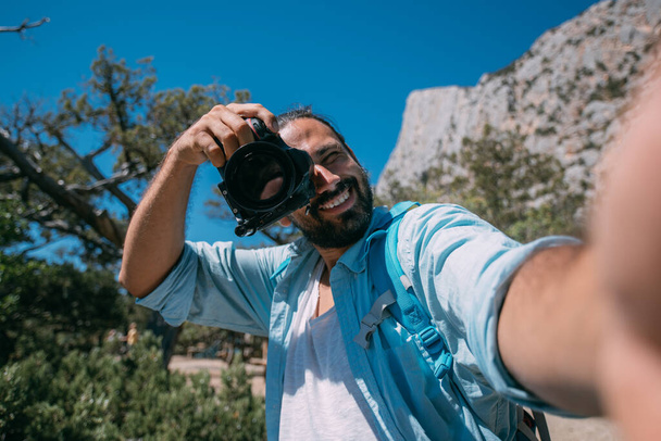 Молодой человек - турист, фотограф улыбается и делает селфи в горах. Красивый парень с рюкзаком и профессиональной камерой сфотографирован рядом с высокими скалами, деревьями и голубым небом - Фото, изображение
