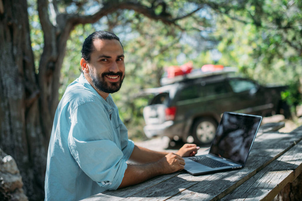 Männliche Touristen arbeiten draußen auf einem Campingplatz an einem Laptop. Junger Mann, Freiberufler, der bei einer Bergwanderung im Freien am Laptop arbeitet. Die Fähigkeit, von jedem Ort der Welt aus zu arbeiten. - Foto, Bild