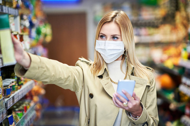 Ενηλίκων γυναίκα σε ιατρική μάσκα χρησιμοποιώντας smartphone και ψώνια για ψώνια - Φωτογραφία, εικόνα
