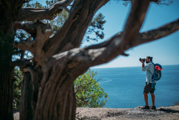 Fotografo turistico di sesso maschile su una scogliera con vista sul mare. Un giovane ragazzo con uno zaino turistico e una macchina fotografica si trova in un punto di vista tra gli alberi e guarda la vista panoramica dell'oceano - Foto, immagini