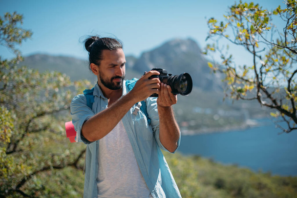 Male tourist, photographer in a mountain grove overlooking the sea. Young handsome guy with tourist backpack and camera on the viewpoint in the trees and looks at the scenic view of the ocean - Фото, изображение