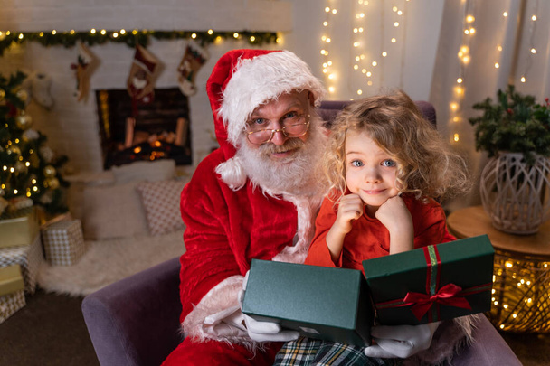 Funny Santa Claus sitting on his rocker with little cute boy sitting on his knee, opening up a gift with something special together. Christmas spirit, holidays and celebrations concept - 写真・画像