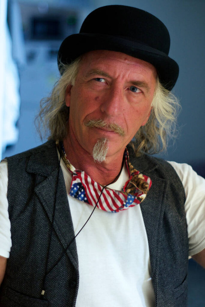 Portrait of handsome blonde, blue-eyed man in bowler hat and vest with american flag coved-19 mask around neck. Looking to the side with long hair, mustache, and goatee. - Photo, Image