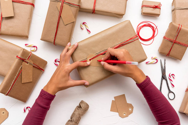 Рука женщины пишет рождественские открытки на подарочных коробках, украшенных крафтовой бумагой - Фото, изображение