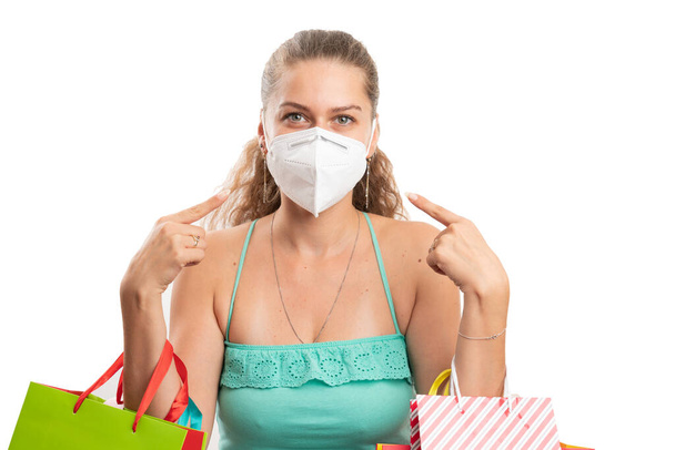 Frau mit Einkaufstaschen, die mit dem Zeigefinger auf chirurgische oder medizinische Einmalmasken zeigen, um eine Kontamination mit dem covid19-Grippevirus zu verhindern, das auf weißem Hintergrund isoliert ist - Foto, Bild