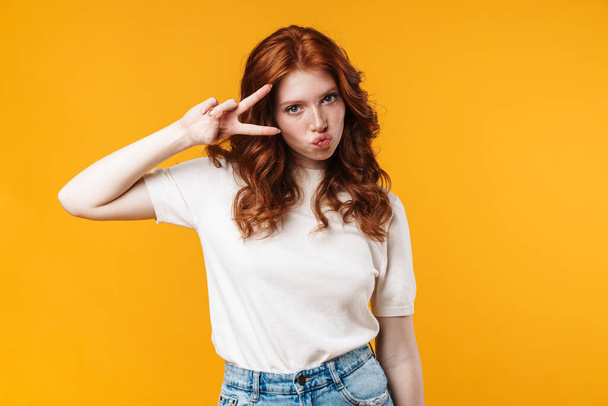 Image of ginger nice girl making kiss lips and gesturing peace sign isolated over yellow background - Fotografie, Obrázek