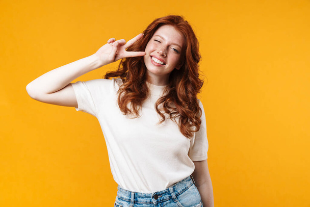 Image of ginger joyful girl winking and gesturing peace sign isolated over yellow background - Foto, Bild