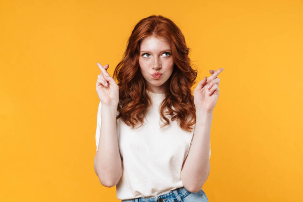 Image of ginger thinking girl holding fingers crossed for good luck isolated over yellow background - Photo, Image