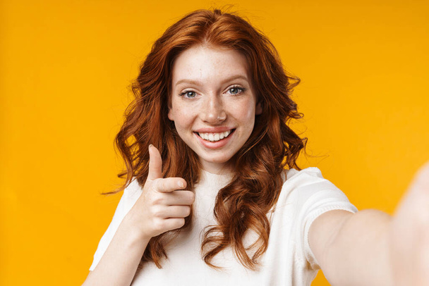 Image of redhead cheerful girl gesturing thumb up and taking selfie photo isolated over yellow background - Photo, image