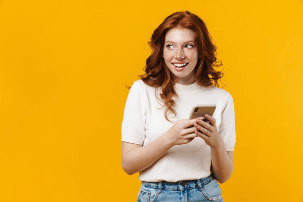 Image of ginger happy girl smiling and using mobile phone isolated over yellow background - Foto, Imagem