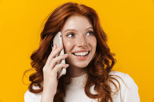 Image of ginger happy girl smiling and talking on mobile phone isolated over yellow background - Zdjęcie, obraz