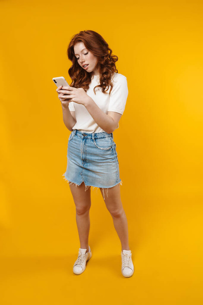 Image of ginger focused girl posing and using mobile phone isolated over yellow background - Foto, Bild