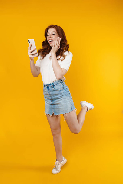 Image of ginger excited girl expressing surprise while using mobile phone isolated over yellow background - Foto, afbeelding