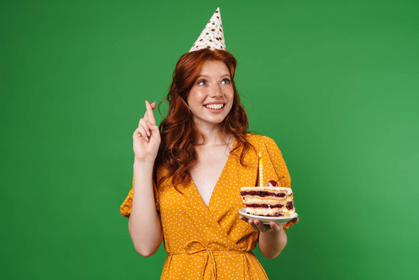 Image of ginger happy girl with fingers crossed holding birthday cake isolated over green background - Valokuva, kuva