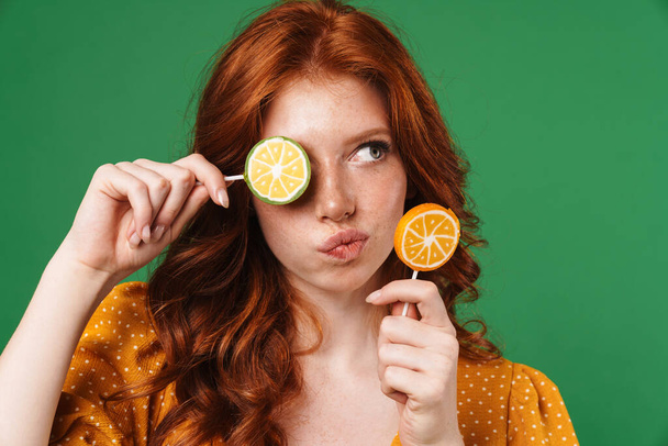 Image of redhead thinking girl posing with lollipops isolated over green background - Фото, изображение