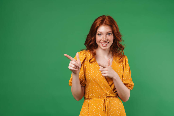 Image of ginger laughing girl pointing fingers and looking aside isolated over green background - Photo, image